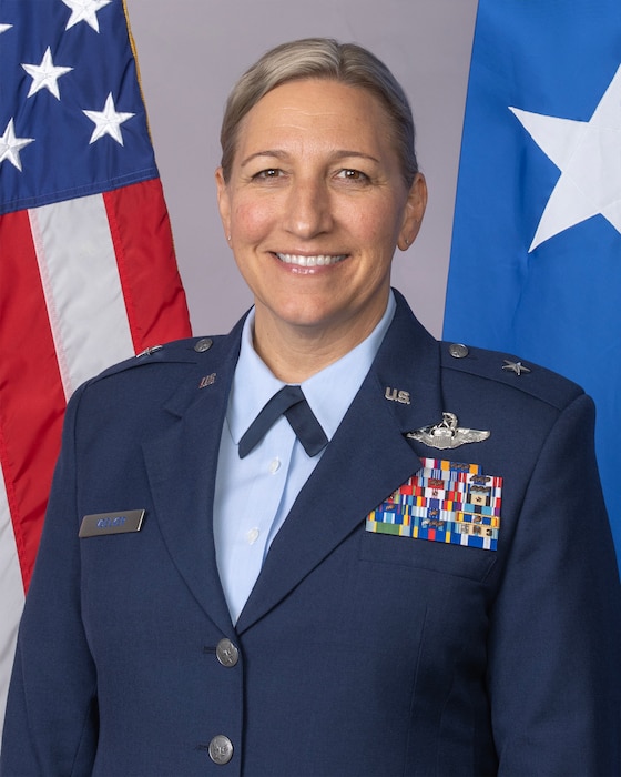 Female general officer photographed in dress blue service uniform, American and One-Star Air Force Flag behind.
