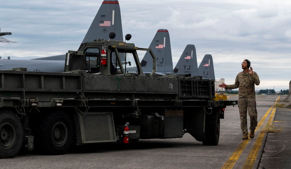 A military member marshals a vehicle being loaded onto an aircraft.