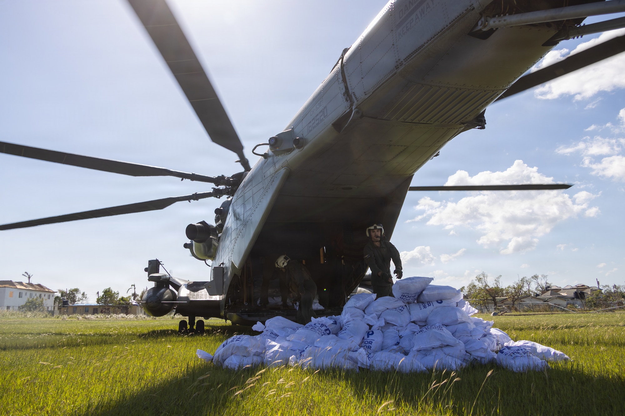 2 people in military uniforms stand outside a helicopter with bags of food in front of them.