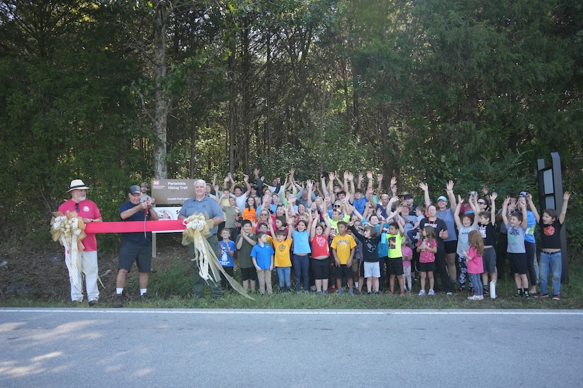 Granville, Tenn. (Sept. 27, 2025) — John Deane (left), owner of Wildwood Resort & Marina, holds one end of the ribbon as Cordell Hull Lake volunteer Ted Tillman cuts it and Kenny Claywell holds the other end during the ribbon-cutting ceremony for the new Storybook Trail at Periwinkle Trail, with volunteers standing to their left. (USACE photo by Noe Gonzalez)