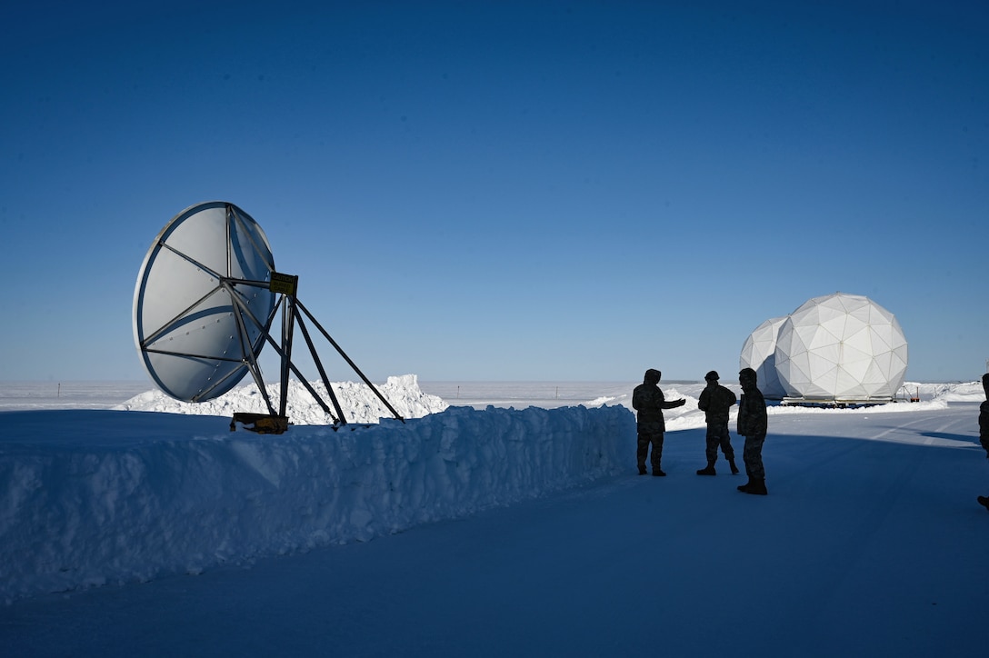 Airmen in silhouette talk in a snowfield with a satellite dish and round white structures in the distance.