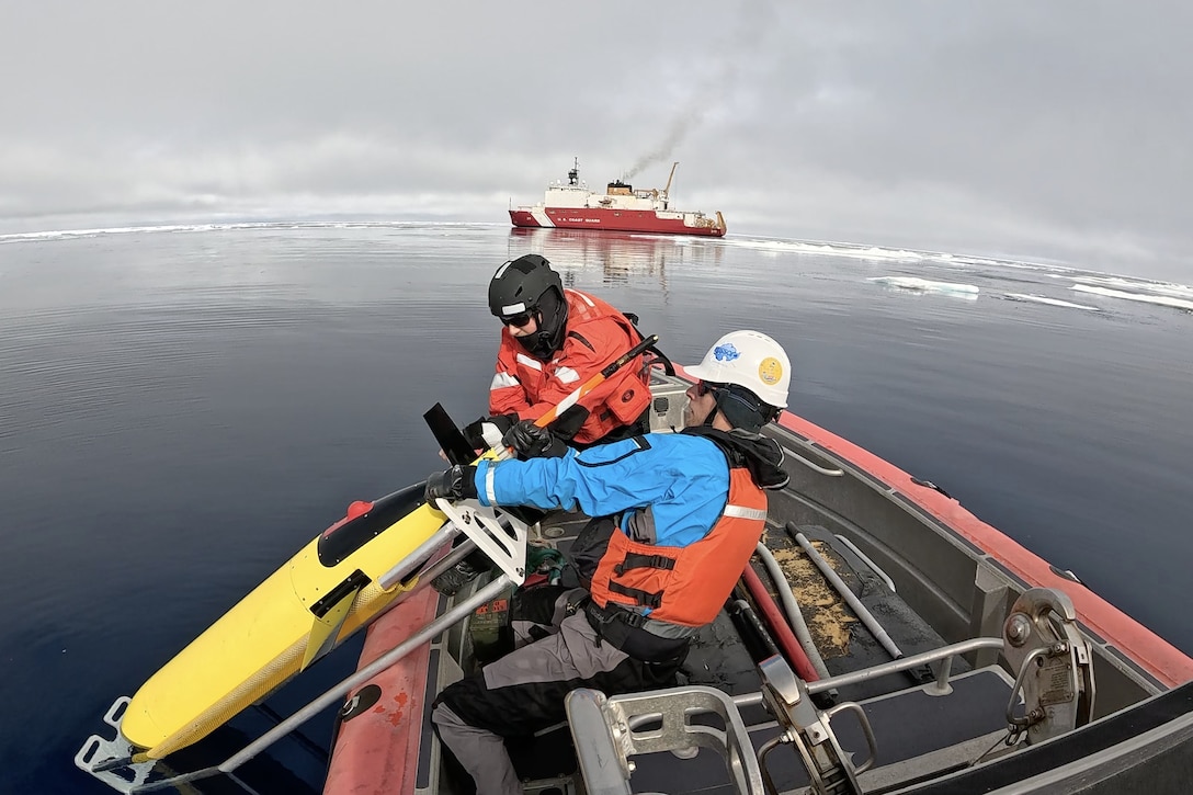 A civilian pulls a yellow craft from water aboard a small boat as a Coast Guardsman looks on and a cutter operates in the distance.