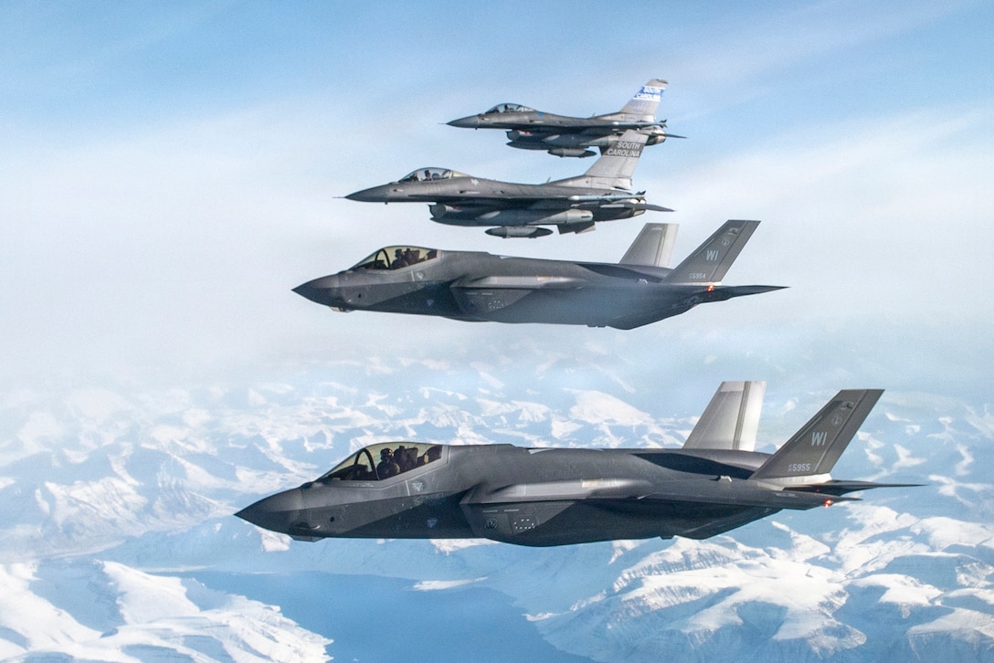 Four Air Force jets fly in cloud-streaked blue sky over snowy terrain.