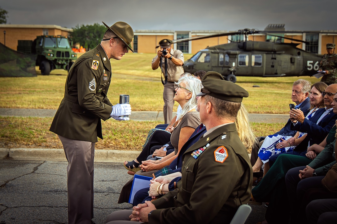 A soldier holds out an artillery shell casing while facing a seated civilian at an outdoor ceremony.