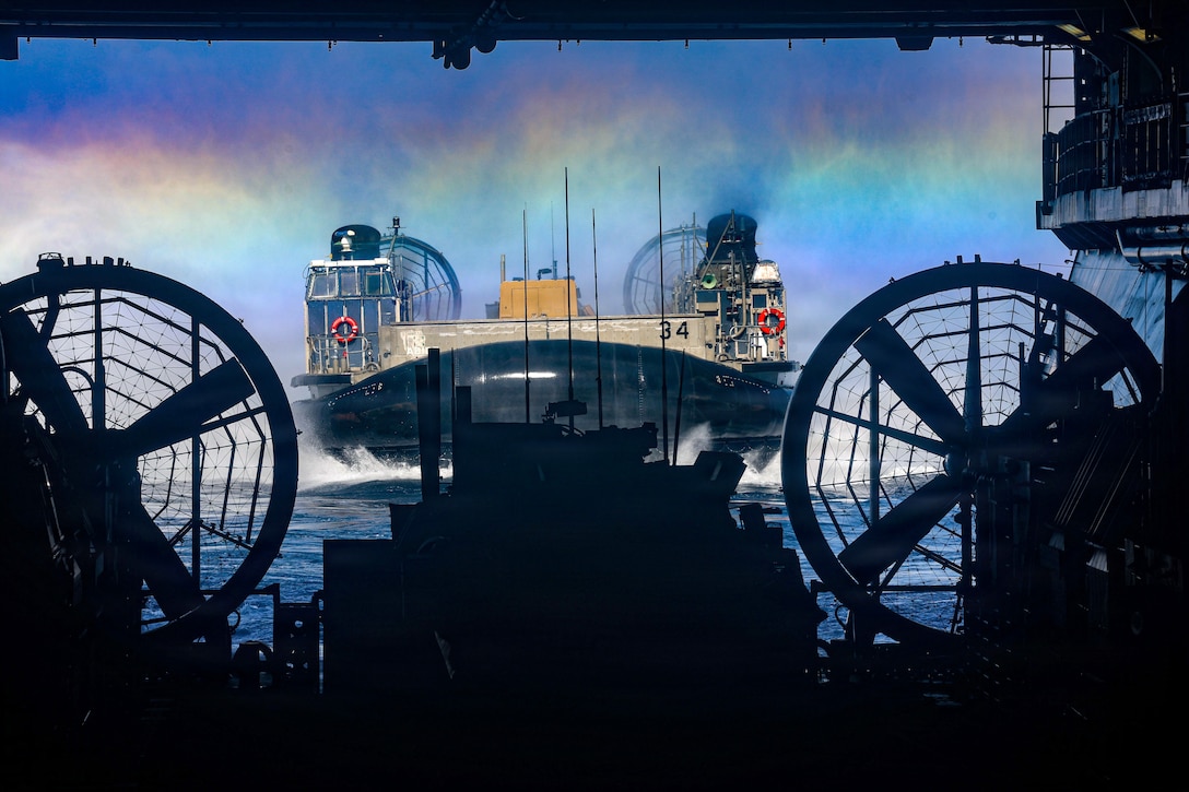 A landing craft approaches a ship's well deck in blue water under a blue sky with a horizontal rainbow.