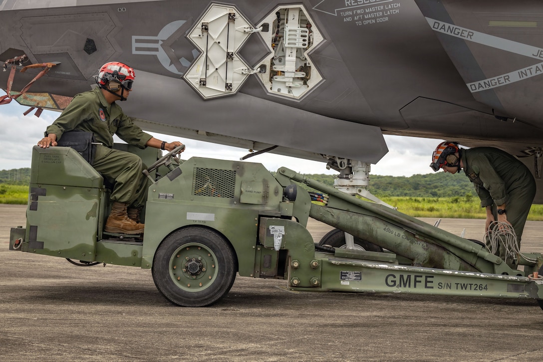 A Marine behind the wheel of a vehicle watches as another stands under an adjacent aircraft parked on a flight line.