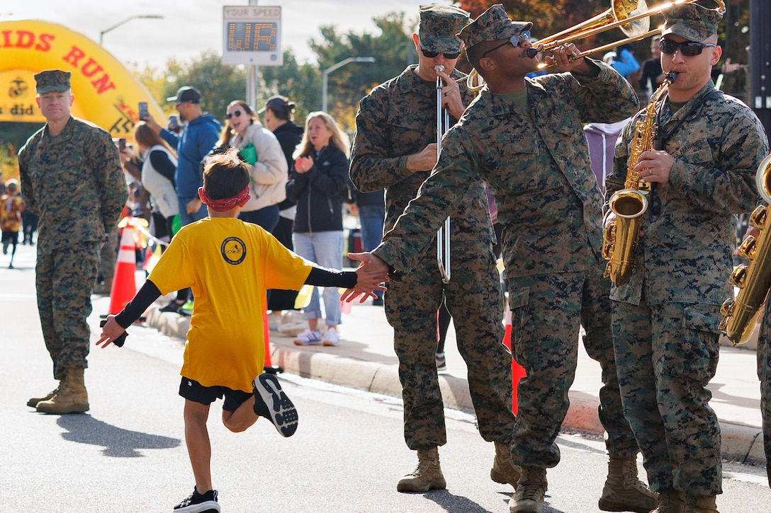 A running child, shown from behind, high-fives a Marine playing trombone with fellow Corps musicians on a roadside.