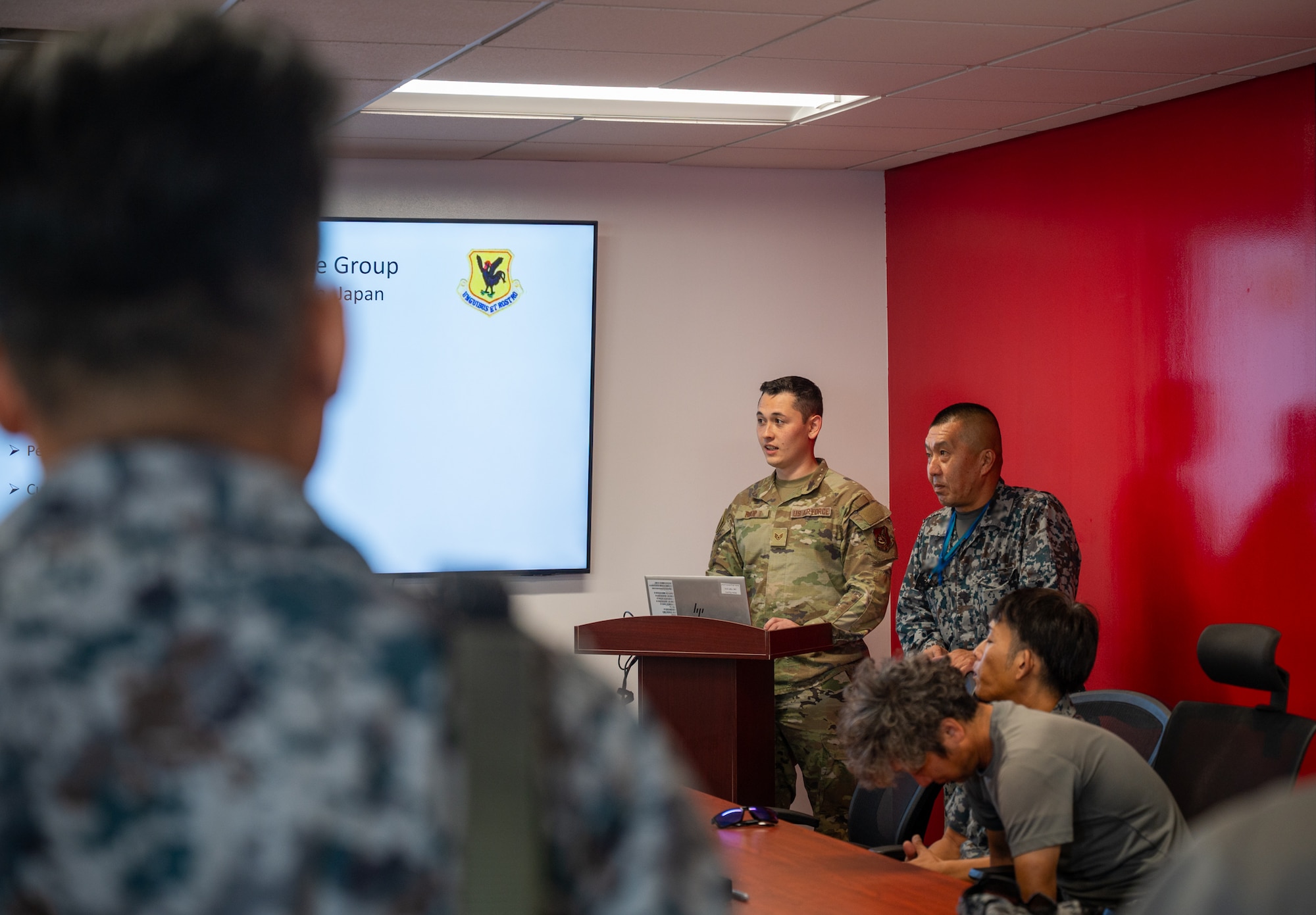 A service member stands a podium and briefs a room full of people.