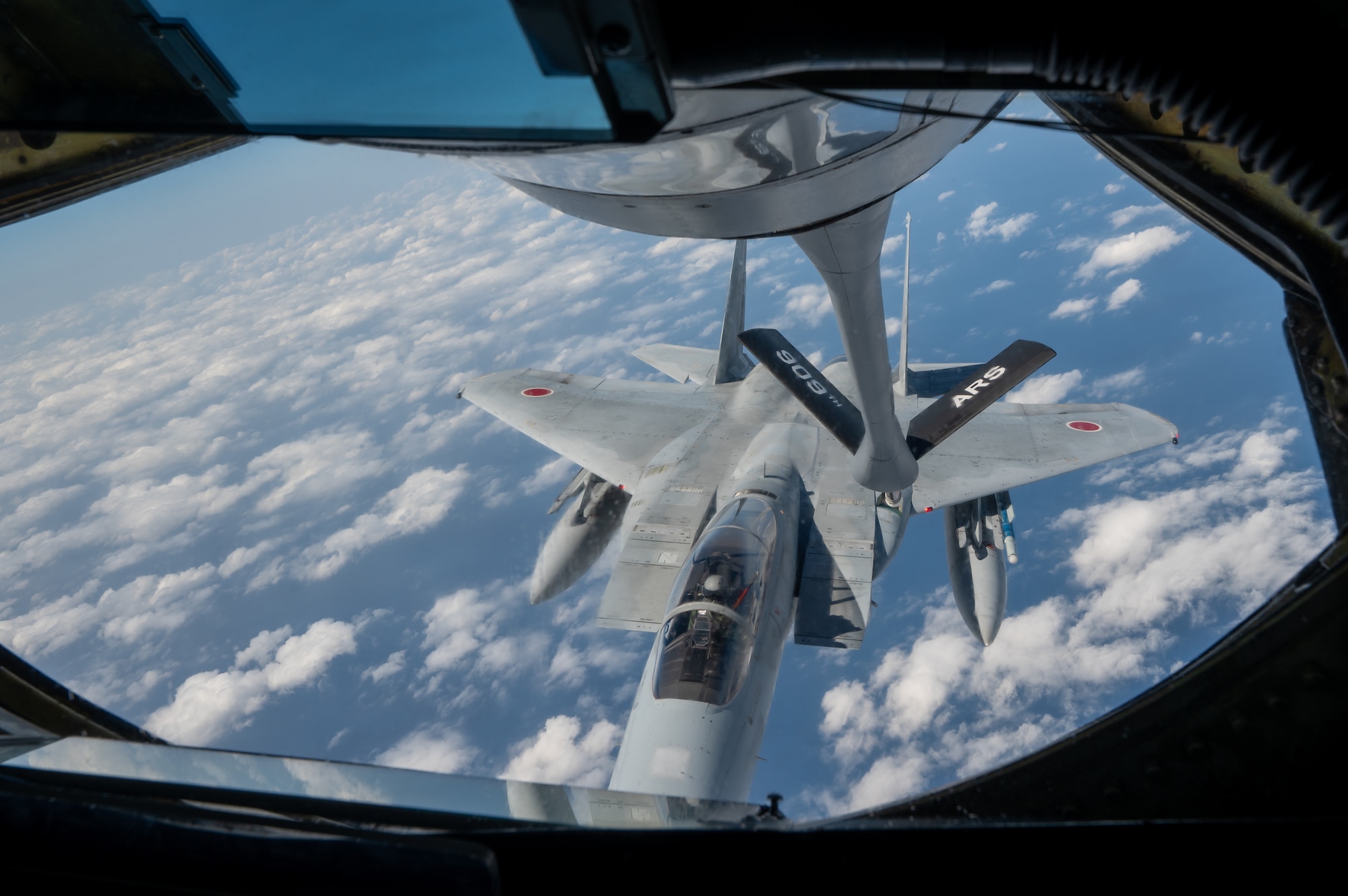 a fighter jet flies amongst clouds nect to a refuel pod