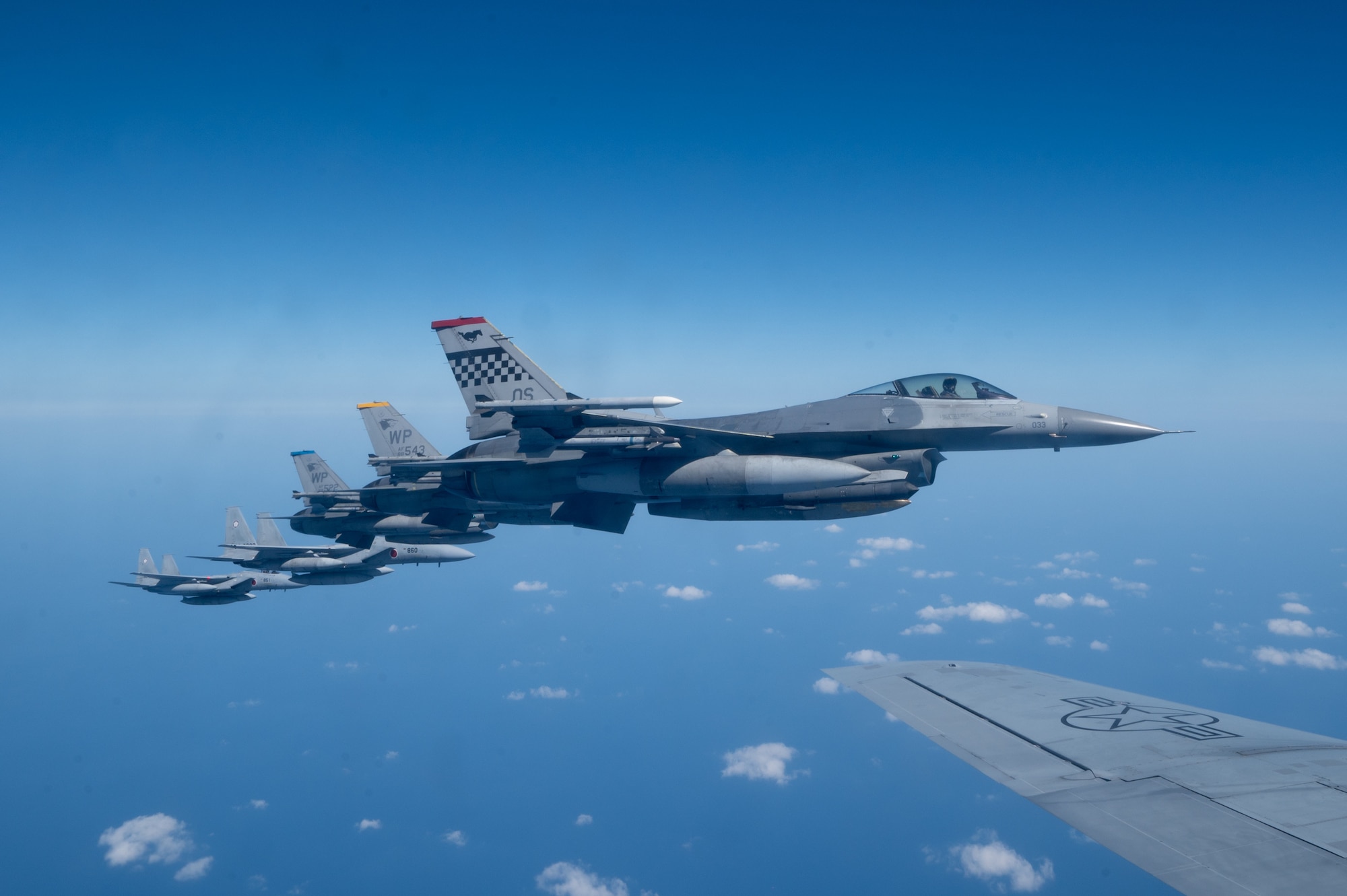 the side view of seven fighter jets in formation flying next to the wing of a KC-135 Stratotanker