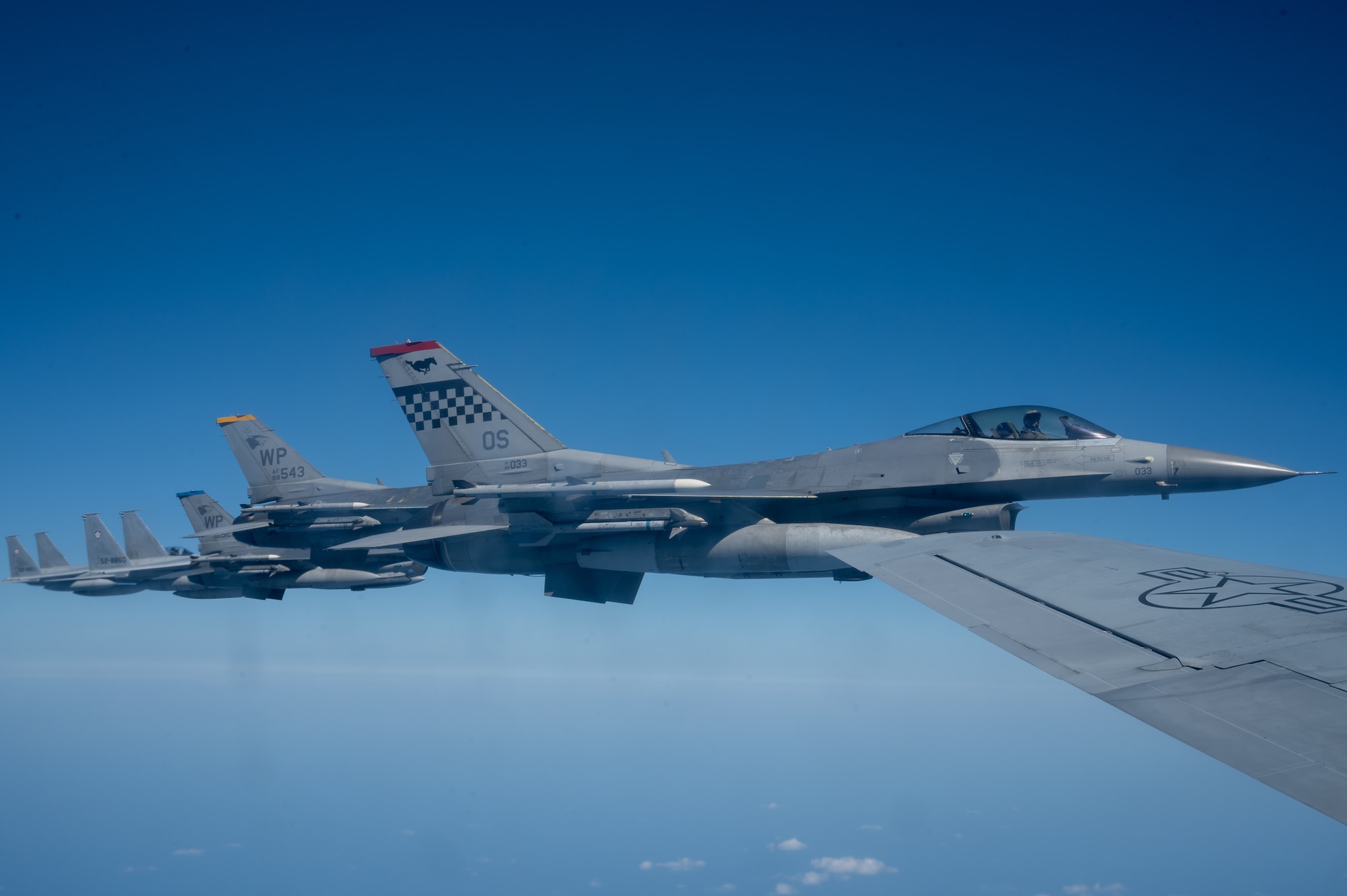 side view of 7 fighters flying in formation next to the wing of a KC-135 stratotanker