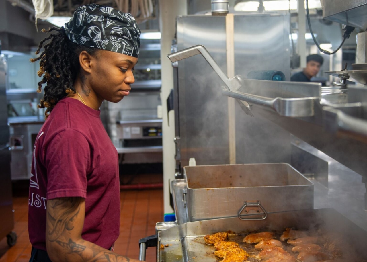 A woman wearing a T-shirt and hat flips meat on a large, steaming grill.