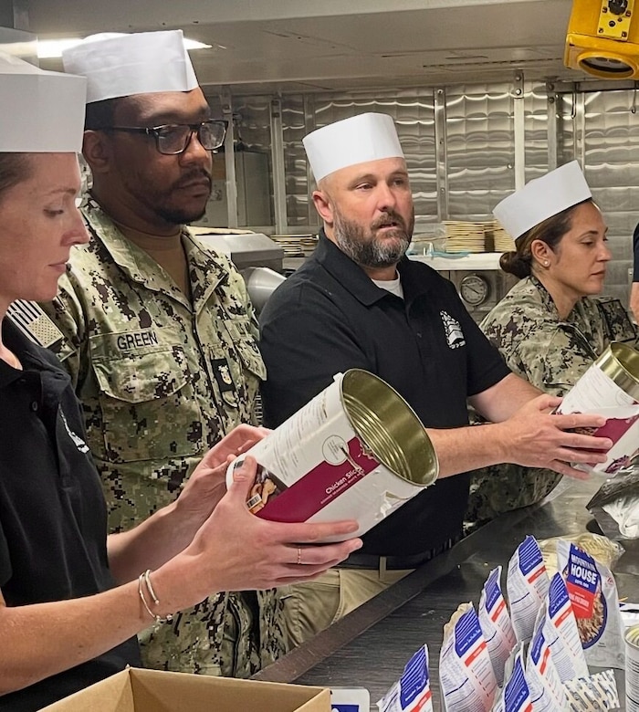 Two people wearing polo shirts and chef hats hold large, opened cans of food as two others in military camouflage uniforms and chef hats look on. Bags of freeze-dried items sit in front of them.