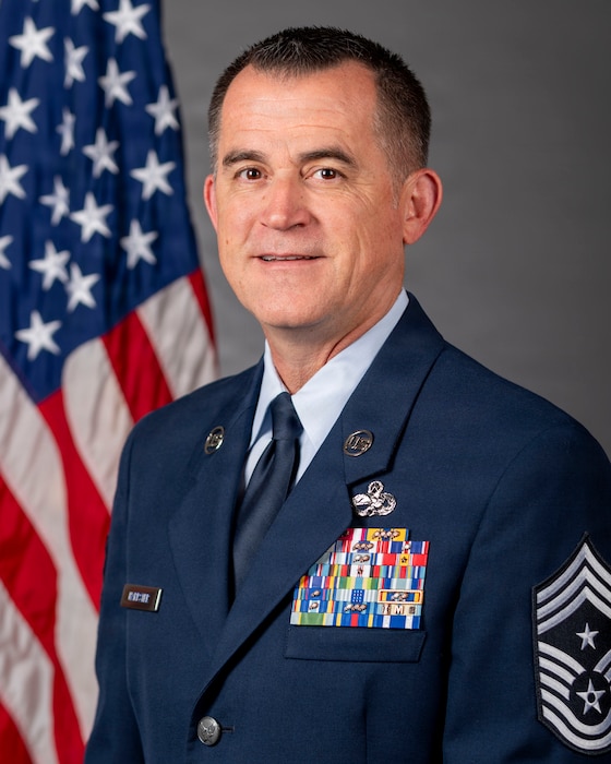 U.S. Airman with Chief Master Sgt Stripes, 5 stripes point down and 3 stripes pointed up with a star between the 3 and 5 stripes indicating a chief Master Sgt. The airman is in front of a U.S. flag in front of a grayish background.