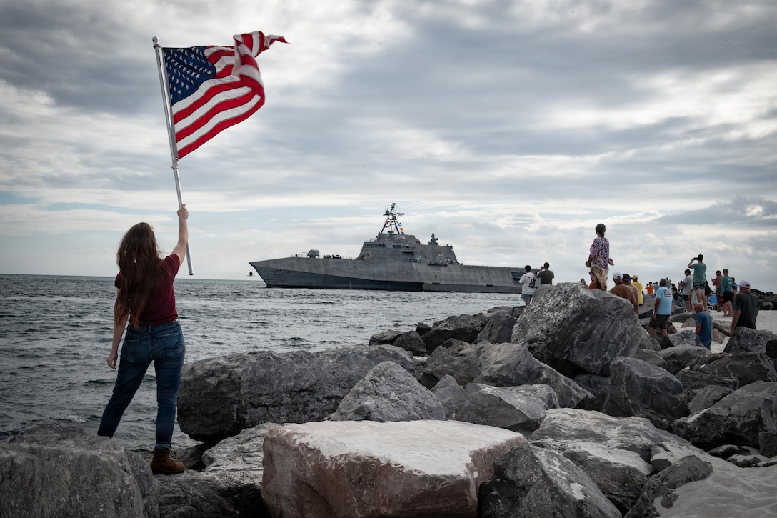 A person holding an American flag and fellow spectators gather on rocks to watch as a ship transits a body of water on a gloomy day.