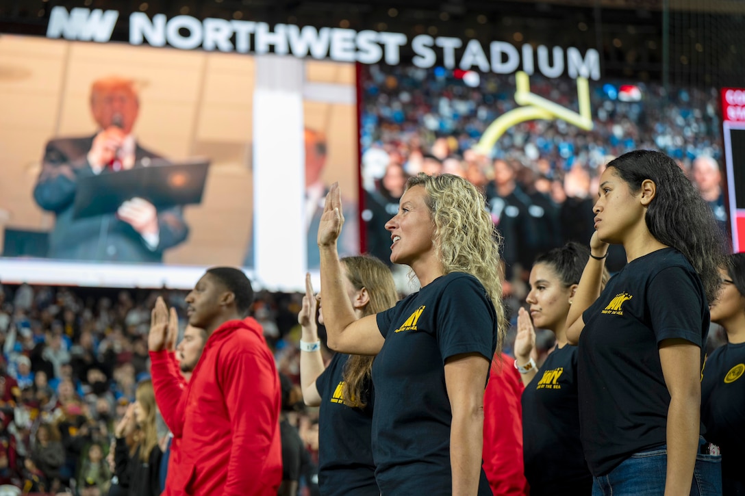 Military recruits raise their right hands as the president reads from a binder as seen on a large screen while fans watch in a stadium.