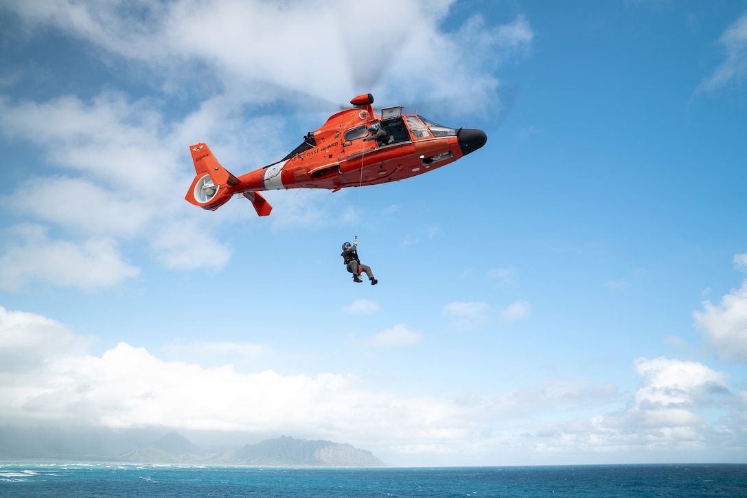 A Coast Guardsman is hoisted to an orange helicopter over a body of water during the day.