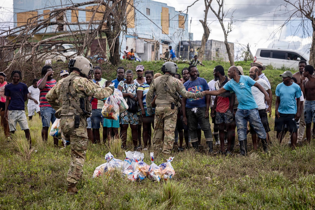 Troops pass out food supplies to dozens of Jamaican citizens in a field with fallen trees and boarded structures in the background.
