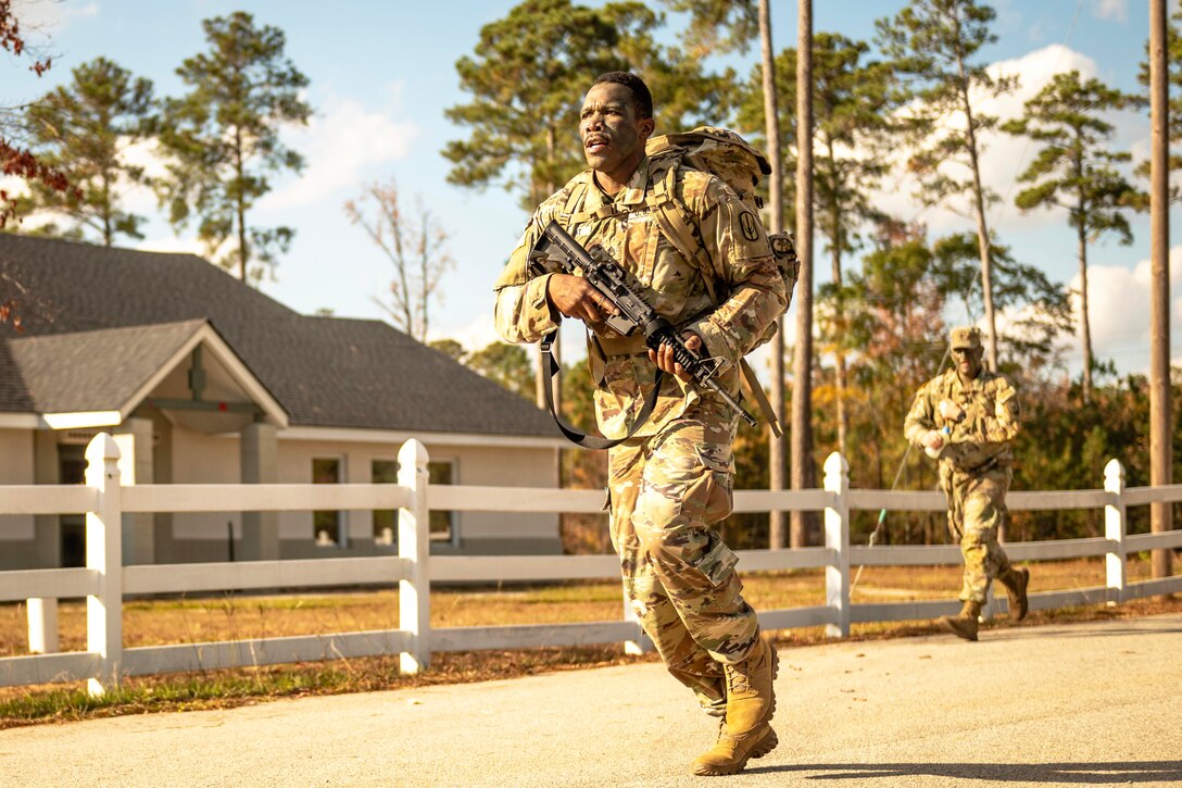 Two soldiers carrying weapons run on a street next to a house on a bright, sunny day.
