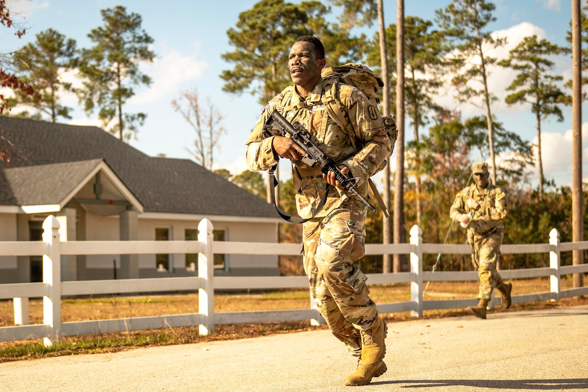 Two soldiers carrying weapons run on a street next to a house on a bright, sunny day.