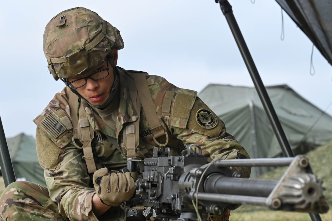 A soldier squats down to load a large weapon with tents in the background under a blue sky.