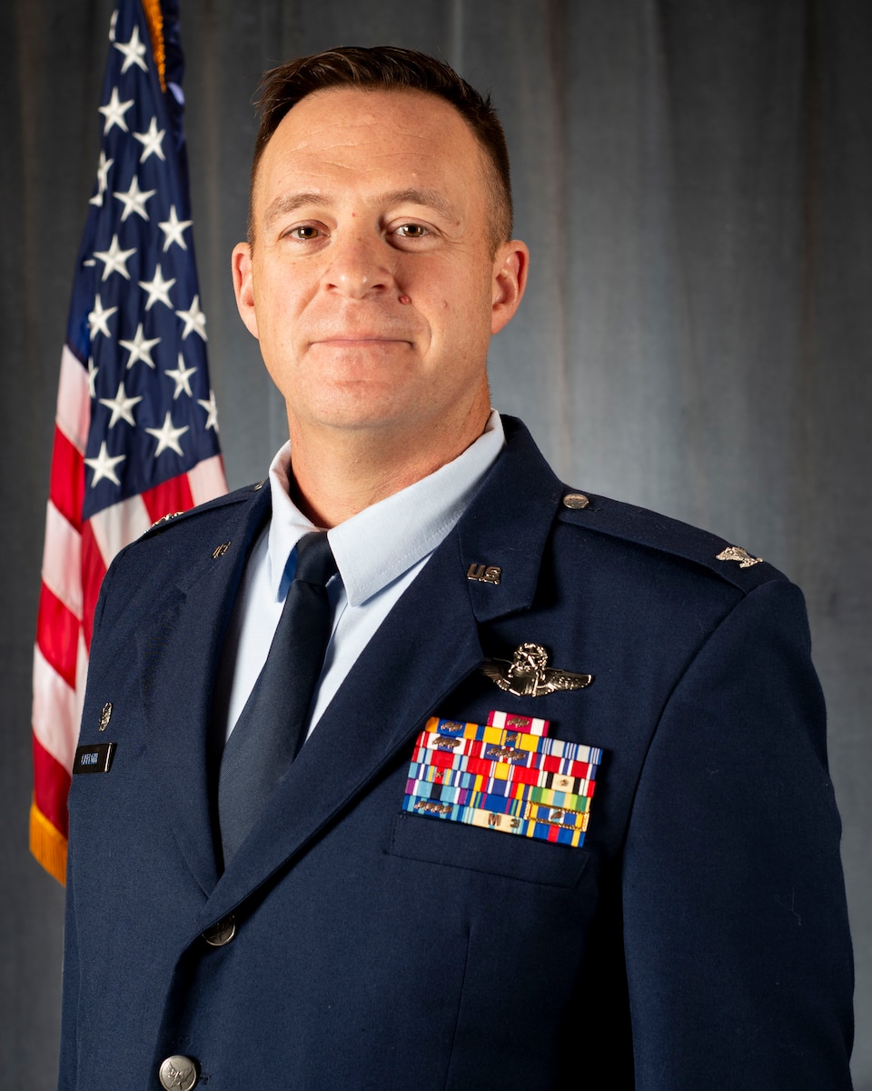 Col. Brian M. Carloni, commander of the 157th Air Refueling Wing, photographed in a studio wearing service-dressed blue uniform standing about 45 degrees of the U.S.A. flag.