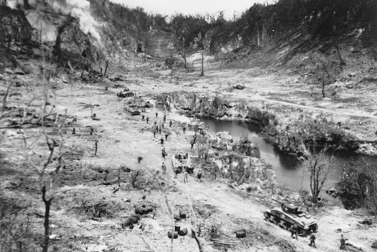 An aerial view of tanks and people walking through a burned-out ravine.
