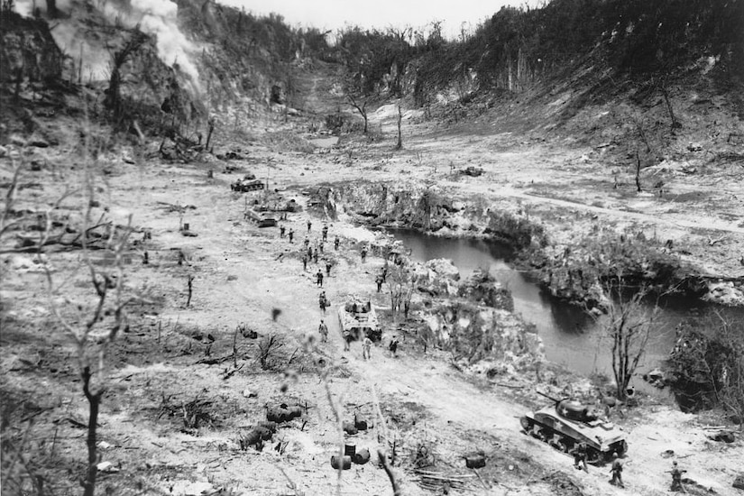 An aerial view of tanks and people walking through a burned-out ravine.