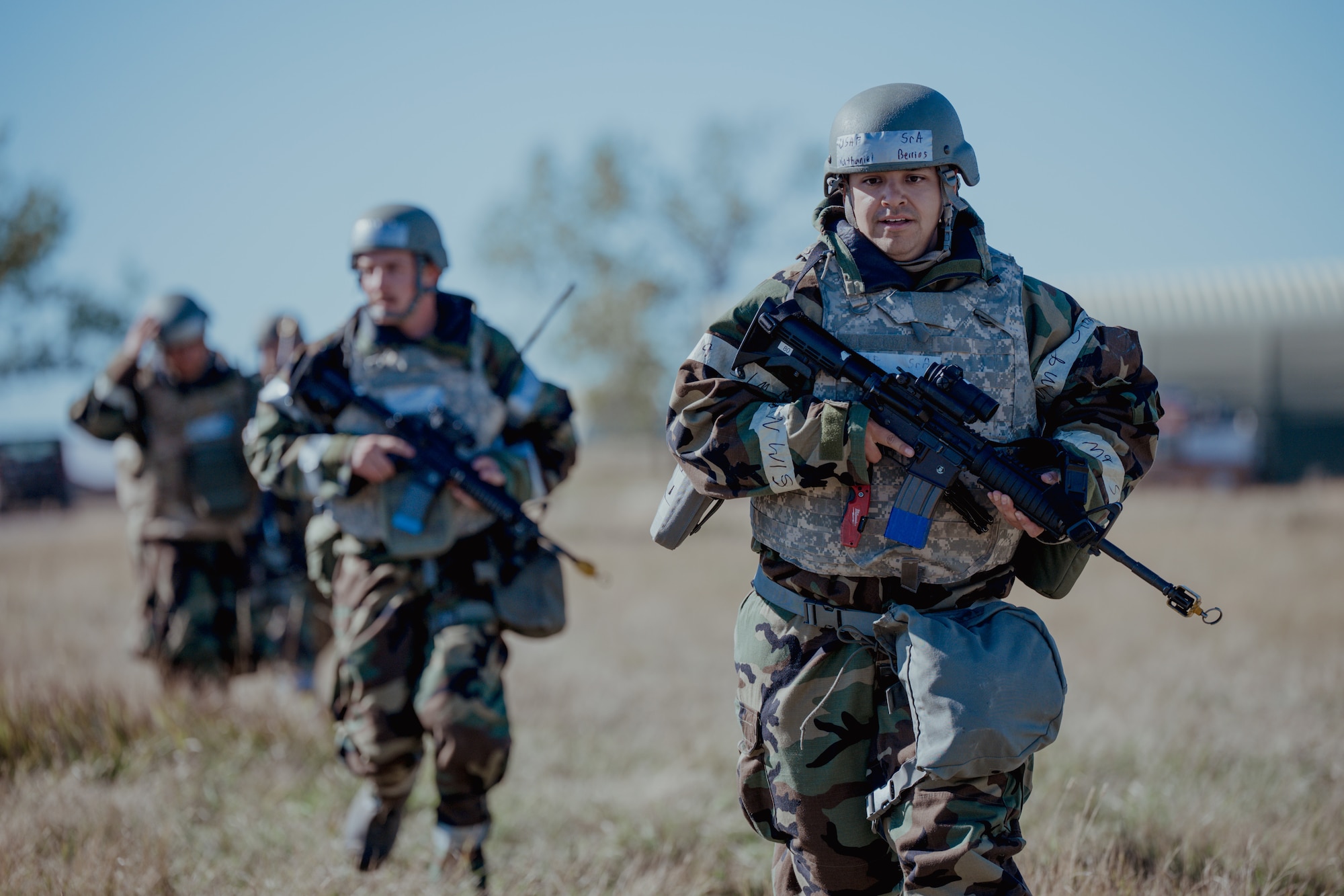 Airmen wearing vests and helmets running towards camera with weapons in hands.