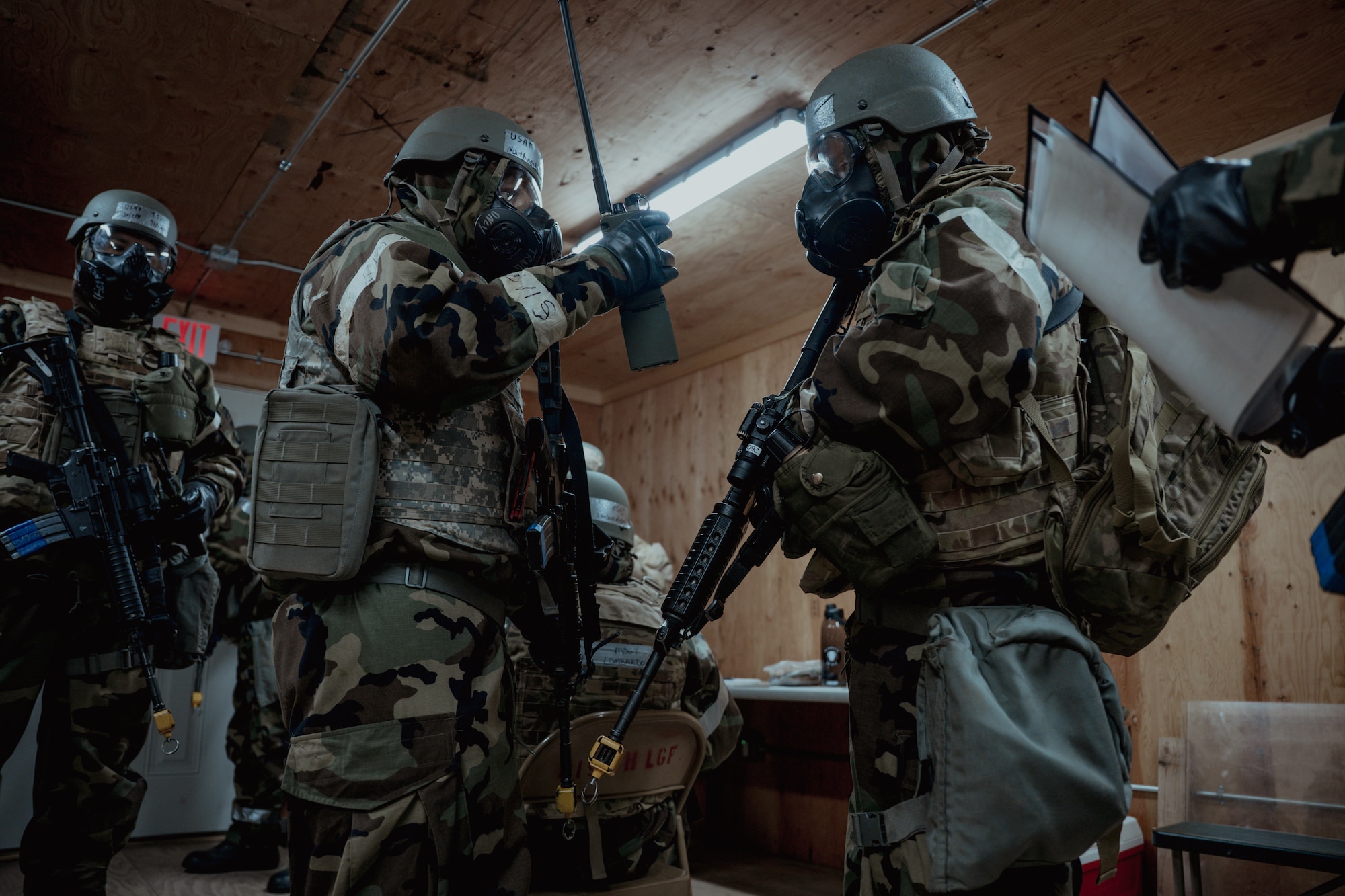 Two Airmen in a room wearing gas masks and helmets communicating with each other.