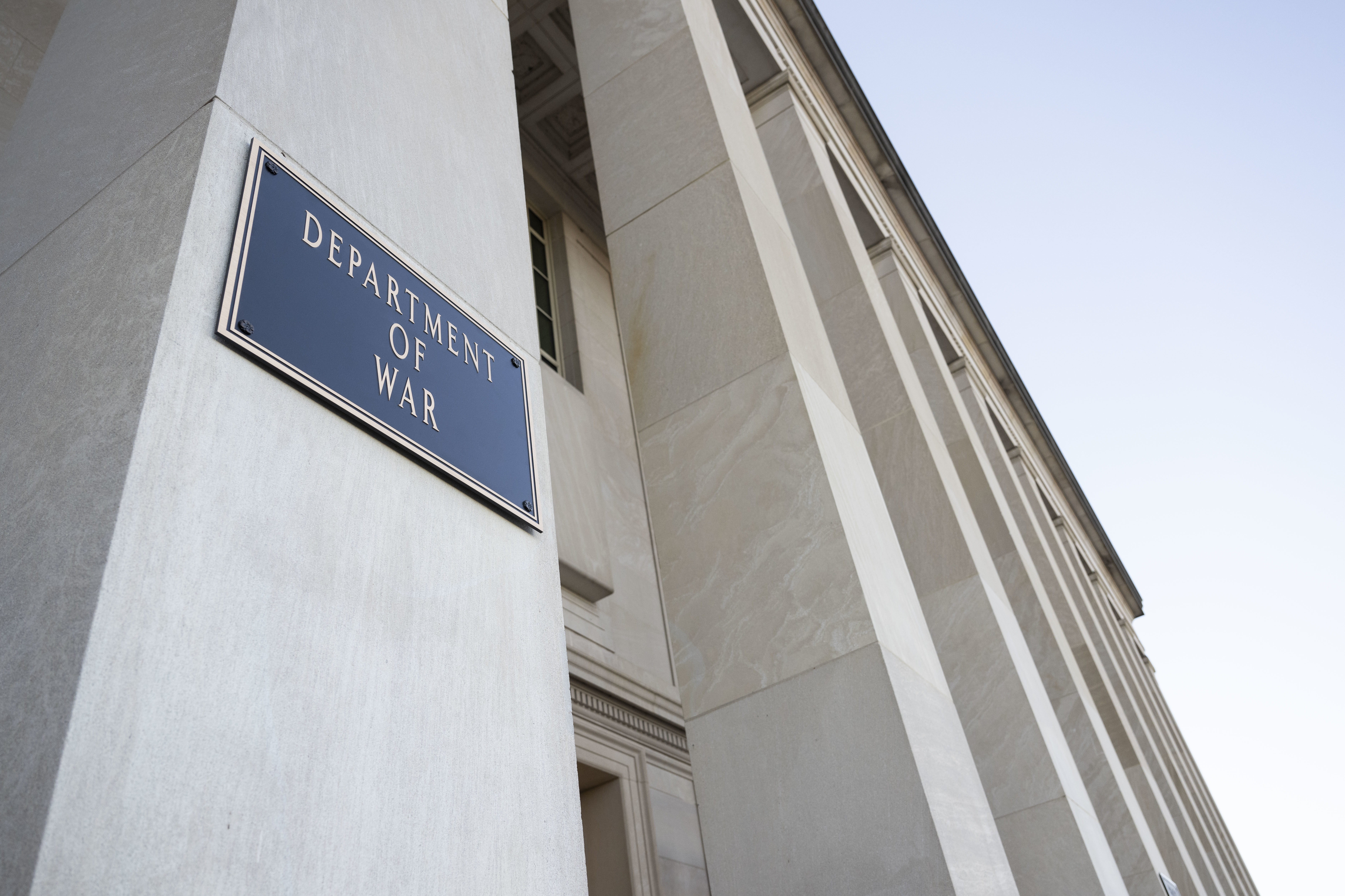 A rectangular metal plaque that reads 'Department of War' is mounted on a large stone column at the entrance of a large building.