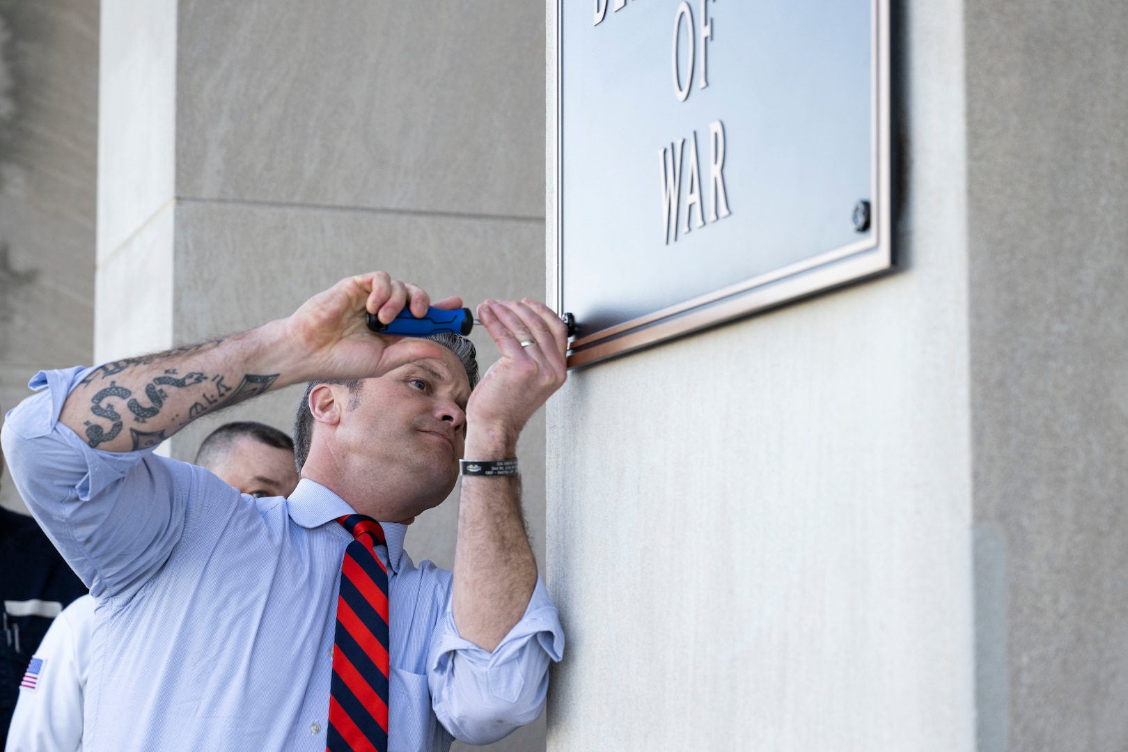 A person in business attire uses a screwdriver to secure a plaque to a concrete wall.