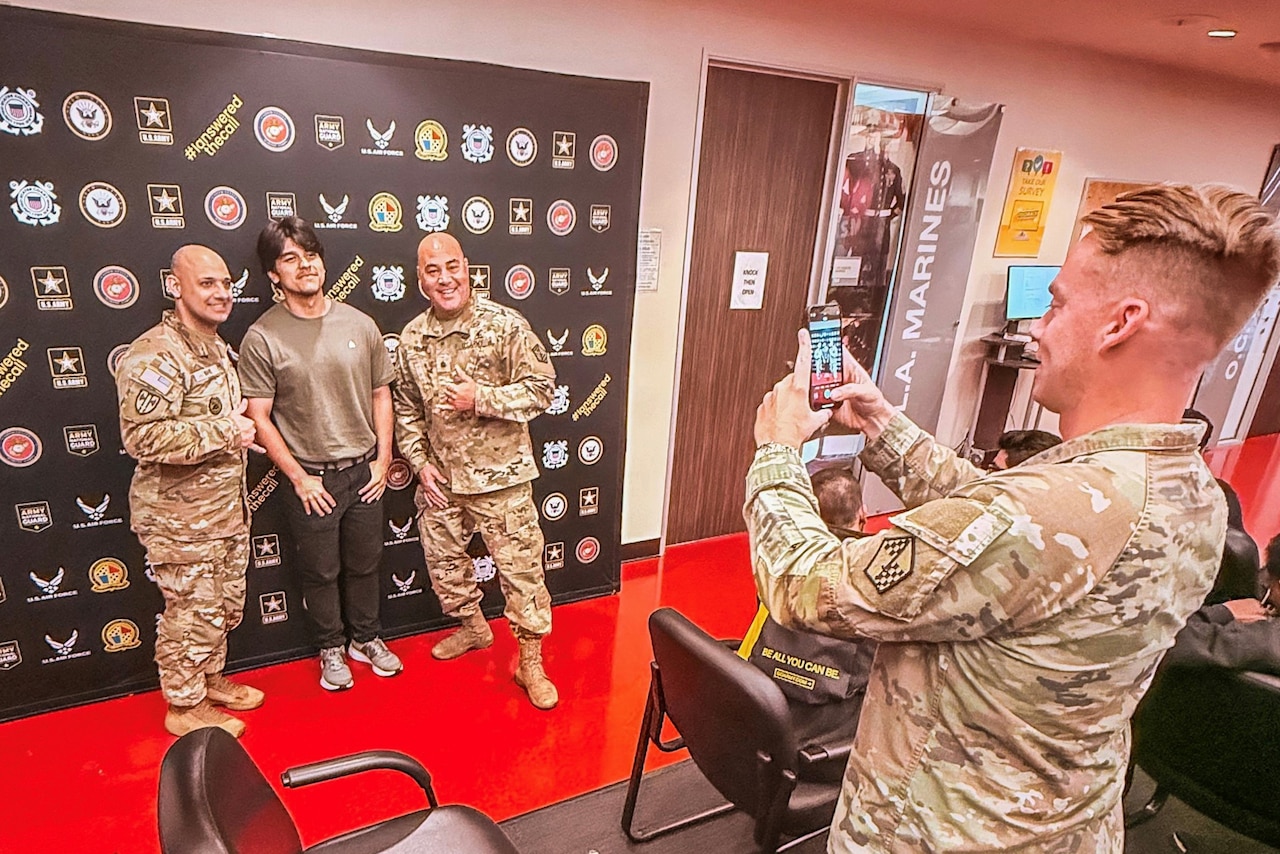 A man wearing a camouflage military uniform holds up a phone as he takes a picture of a man in casual attire and two men in camouflage military uniforms standing in front of a black backdrop with military logos.
