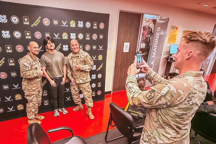 A man wearing a camouflage military uniform holds up a phone as he takes a picture of a man in casual attire and two men in camouflage military uniforms standing in front of a black backdrop with military logos.