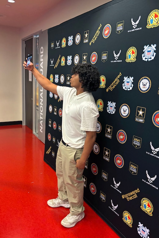 A man in casual attire holds up his phone to take a picture of himself in front of a black backdrop with military logos.