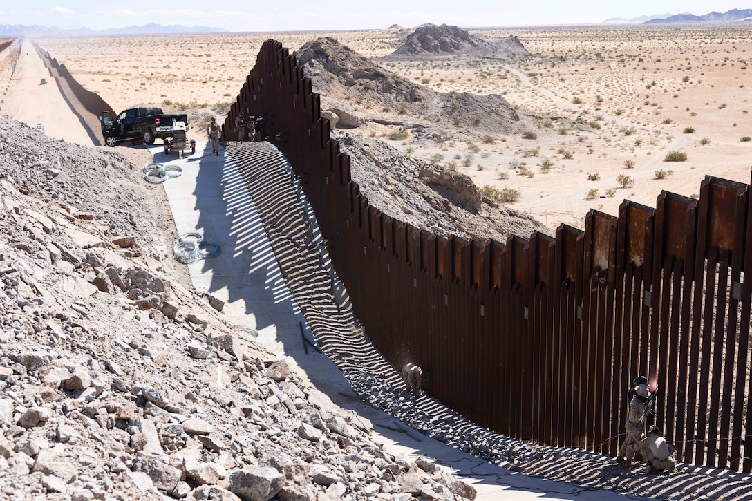Marines work on a large metal fence in a desert-like area on a sunny day.