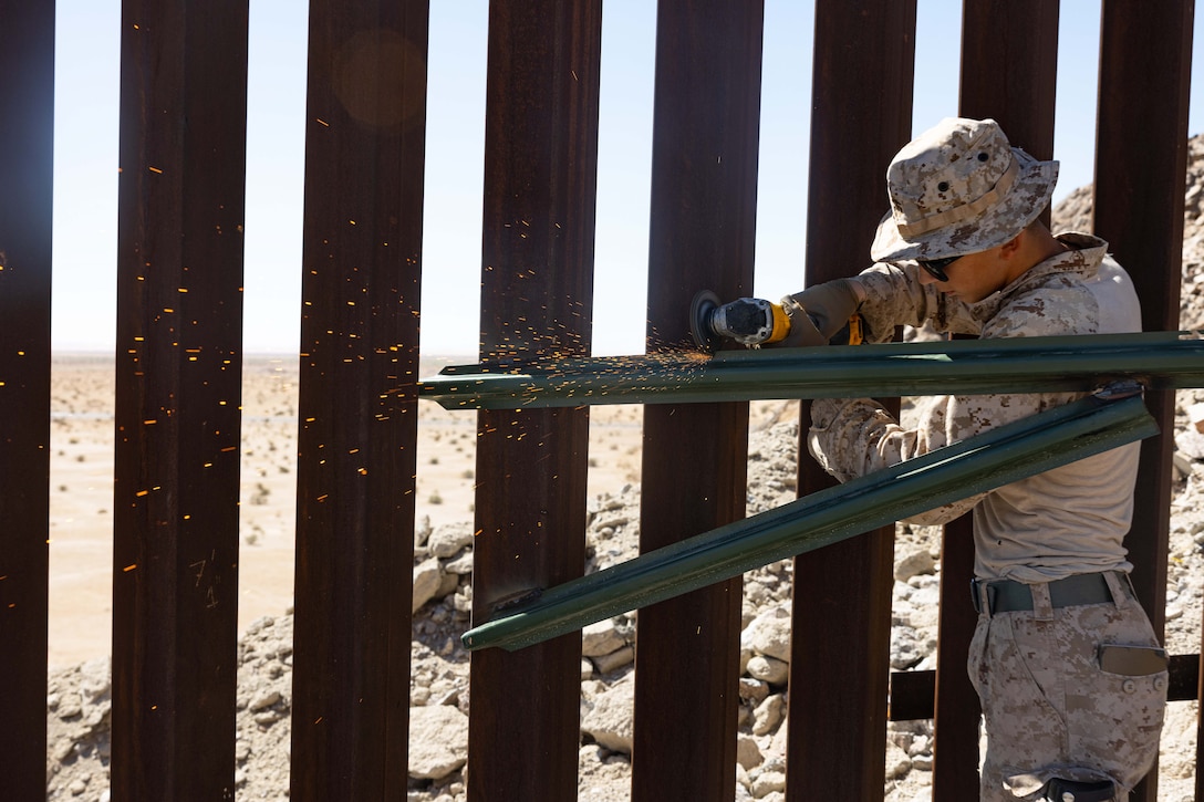 Sparks fly as a Marine welds a bracket for barbed wire attachment in a desert-like area on a sunny day.