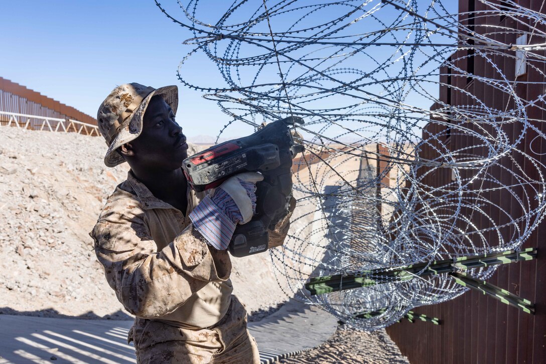 A Marine works on concertina wire attached to a large fence in a desert-like area.