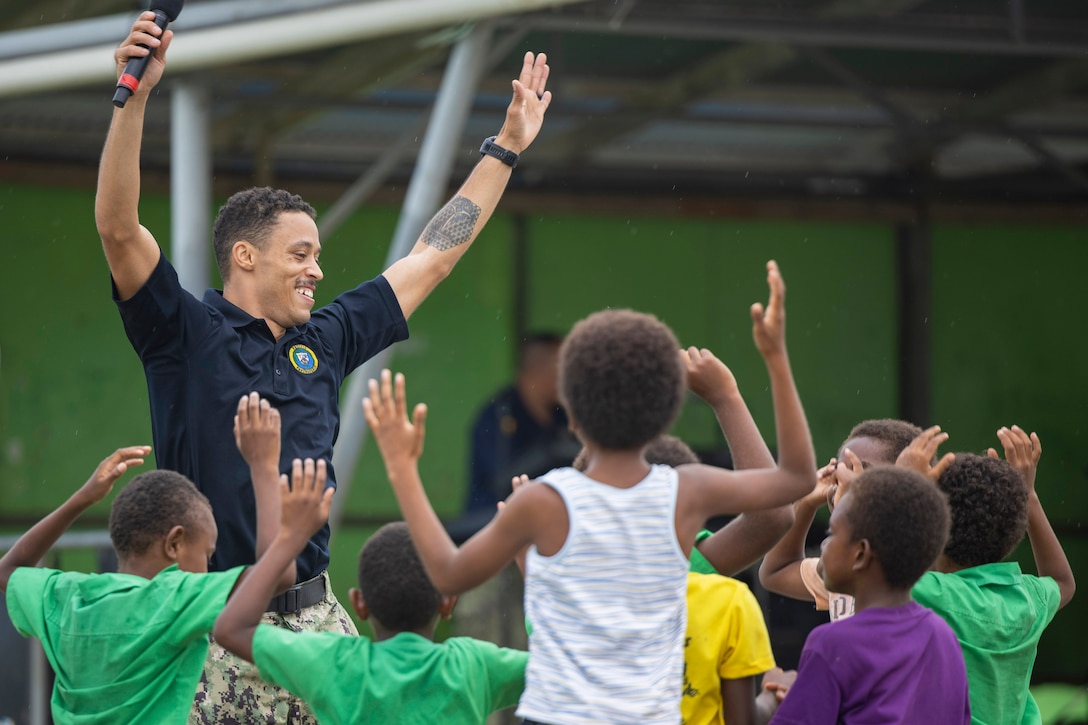 A standing person holding a microphone in one hand with both arms raised smiles at surrounding children, who also are raising their arms outdoors in daytime; a structure and another person are in the background.