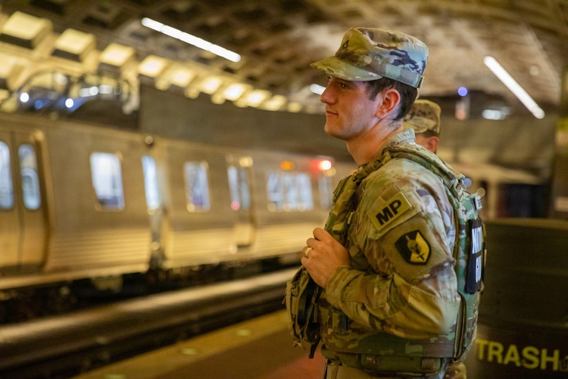 A man dressed in a camouflage military uniform stands on a subway platform.