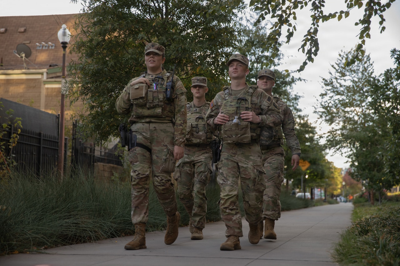 Four soldiers wearing camouflage military uniforms walk down a sidewalk flanked by trees and greenery.