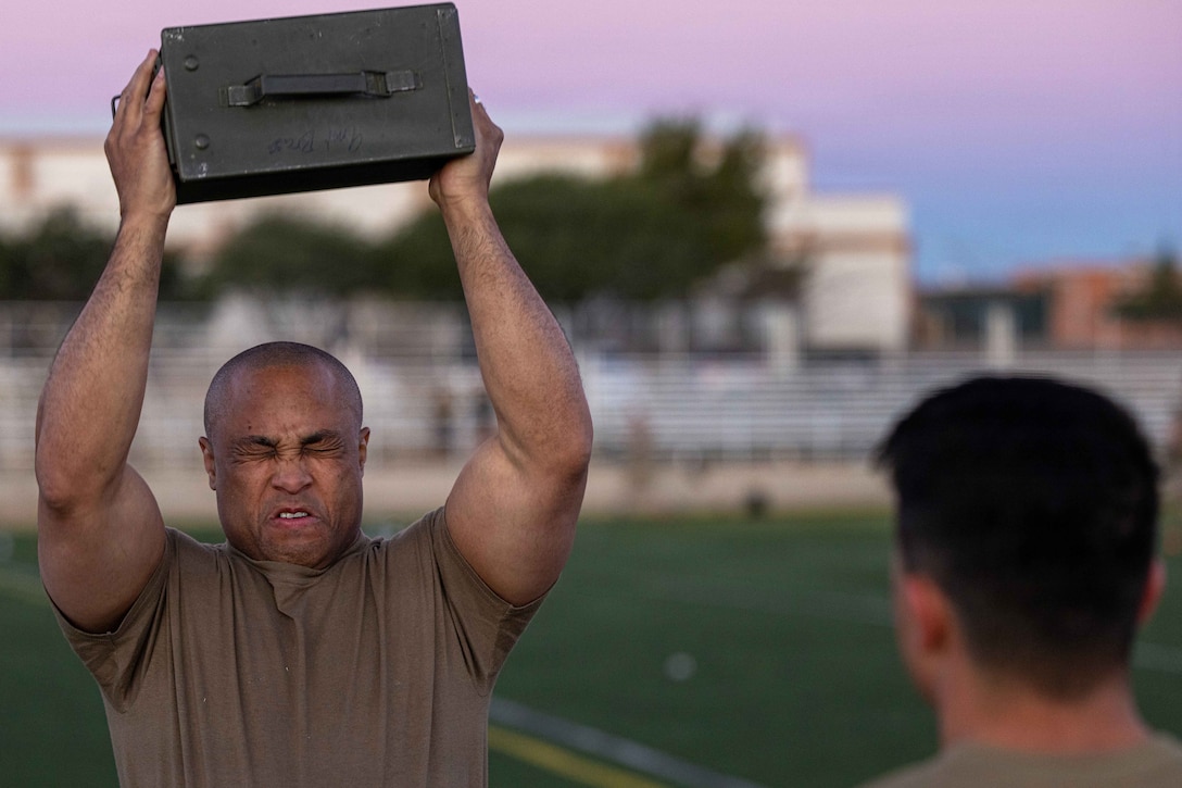 A soldier wearing a green T-shirt grimaces while lifting a cannister with both hands above the head on an outdoor field in daytime, as another person watches nearby.