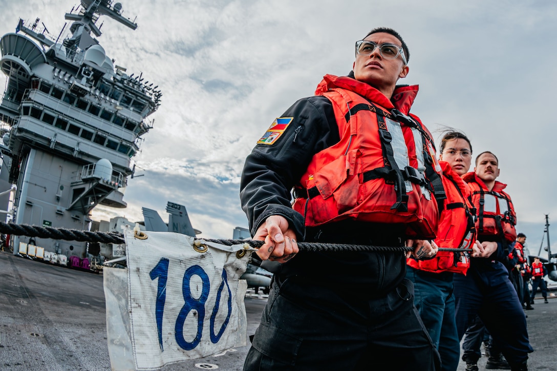 Sailors wearing life vests pull on a line with a small sign reading "180" while standing on the deck of a large ship on a cloudy day, with the control tower, aircraft and other sailors in the background.