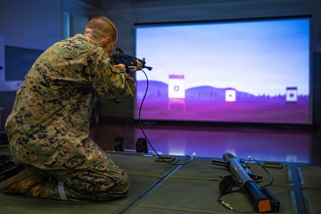 A kneeling Marine in a camouflage uniform aims a rifle at targets on a large screen indoors, with other equipment nearby.