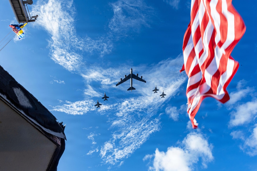 A large aircraft and four fighter jets fly in formation over a ship during daytime, with part of an American flag also visible.