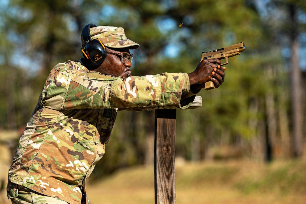 An airman wearing a camouflage uniform and ear protection fires a pistol near a wood post during daytime, with trees in the background.