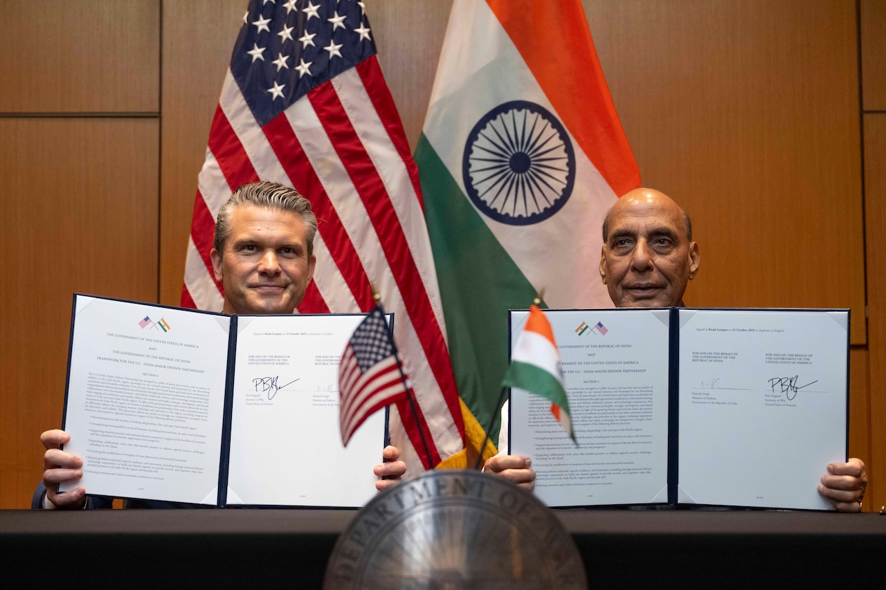 Two men hold up folders containing signed documents. Behind them are the flags of the U.S. and India.