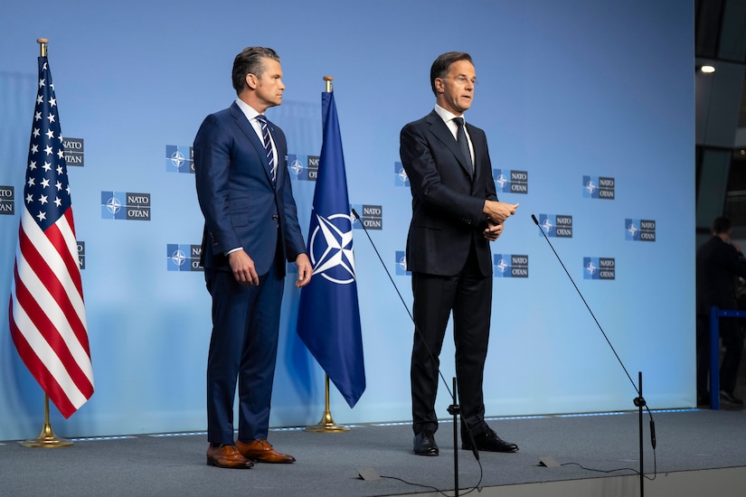 Two men stand together on a stage with the NATO flag and the American flag behind them.
