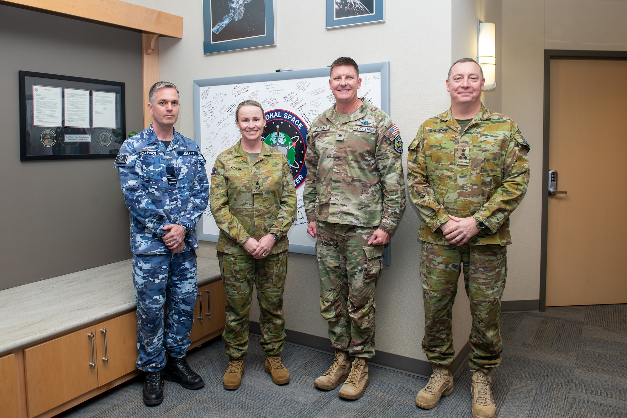 Four uniformed service members stand together, smiling, in front of a whiteboard with signatures on it.