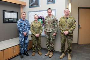 Four uniformed service members stand together, smiling, in front of a whiteboard with signatures on it.